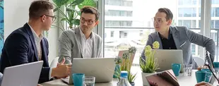 Drei Männer in Business-Kleidung diskutieren an einem Tisch mit Laptops, Pflanzen und Kaffeetassen. Im Hintergrund sind große Fenster mit Blick auf ein Bürogebäude.