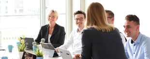 Fünf Personen in Business-Kleidung sitzen in einem modernen, hellen Büro und führen ein Meeting. Sie lachen und arbeiten mit Laptops und Notizen.