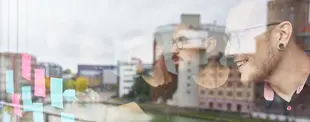 Zwei junge Männer brainstormen vor einer Glaswand mit bunten Klebezetteln. Ihre Reflexionen sind im Fenster zu sehen, mit Stadtgebäuden im Hintergrund.