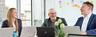 Drei Geschäftsleute in einem modernen Büro lachen während eines Meetings mit Laptops. Im Hintergrund ein Whiteboard mit bunten Notizen.